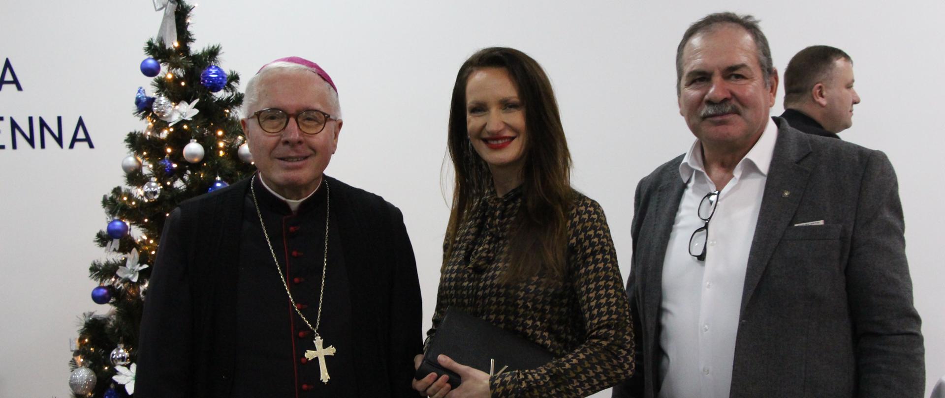 Zdjęcie przedstawia trzy uśmiechnięte osoby stojące obok choinki. Po lewej stronie stoi Ks Biskup Tadeusz Bronakowski. W środku Pani Edyta Marchelska-Groszfeld Starosta Zambrowski, a po prawej mężczyzna w białej koszuli i szarej marynarce z okularami zawieszonymi u dekoldu.