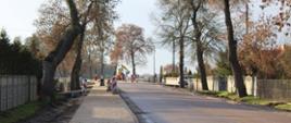 Zdjęcie przedstawia fragment wykonanego ciągu pieszo rowerowego przy drodze powiatowej. W tle robotnicy kontynuujący prace drogowe.