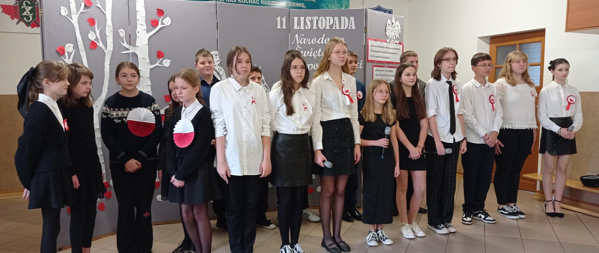 Grupa uczniów ubrana w galowe stroje. Z tyłu za nimi stoi szara ścianka dekoracyjna z napisem 11 LISTOPADA.