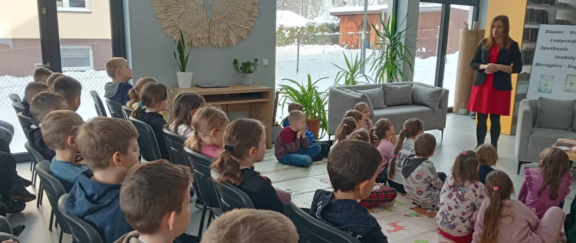 Dzieci siedzą na krzesłach i na podłodzie i słuchają opowieści pani pisarki, w tle regały biblioteczne z książkami