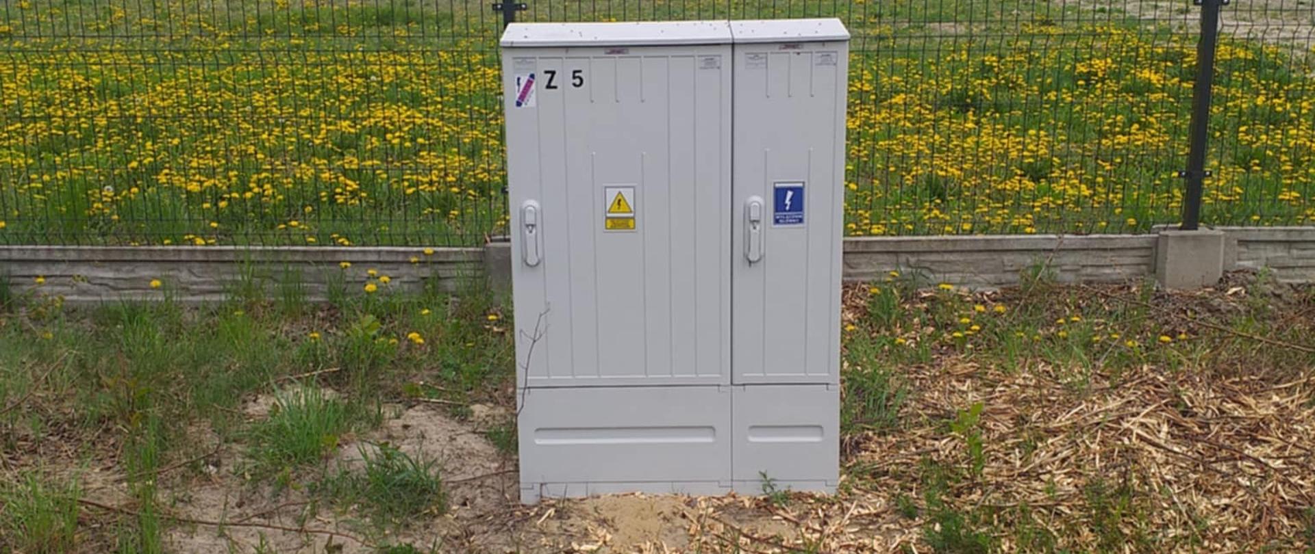 Na pierwszym planie widoczna jest metalowa skrzynka elektryczna w kolorze szarym, stojąca na otwartym terenie pokrytym trawą i mleczami. Skrzynka jest umieszczona przed ogrodzeniem z czarnej metalowej siatki, które oddziela ją od większej przestrzeni. Na skrzynce znajdują się różne oznaczenia, w tym żółty trójkąt z symbolem pioruna ostrzegającym przed wysokim napięciem, niebieska etykieta z białym symbolem oraz numer „Z 5” umieszczony w górnym lewym rogu.
W tle widać drzewa i inne rośliny, a nad nimi rozciąga się zachmurzone niebo, co nadaje obrazowi ponury lub pochmurny nastrój. Całość przedstawia elementy infrastruktury elektrycznej, które są kluczowe dla dostarczania energii do pobliskich obszarów.
