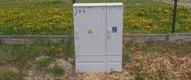 Na pierwszym planie widoczna jest metalowa skrzynka elektryczna w kolorze szarym, stojąca na otwartym terenie pokrytym trawą i mleczami. Skrzynka jest umieszczona przed ogrodzeniem z czarnej metalowej siatki, które oddziela ją od większej przestrzeni. Na skrzynce znajdują się różne oznaczenia, w tym żółty trójkąt z symbolem pioruna ostrzegającym przed wysokim napięciem, niebieska etykieta z białym symbolem oraz numer „Z 5” umieszczony w górnym lewym rogu.
W tle widać drzewa i inne rośliny, a nad nimi rozciąga się zachmurzone niebo, co nadaje obrazowi ponury lub pochmurny nastrój. Całość przedstawia elementy infrastruktury elektrycznej, które są kluczowe dla dostarczania energii do pobliskich obszarów.