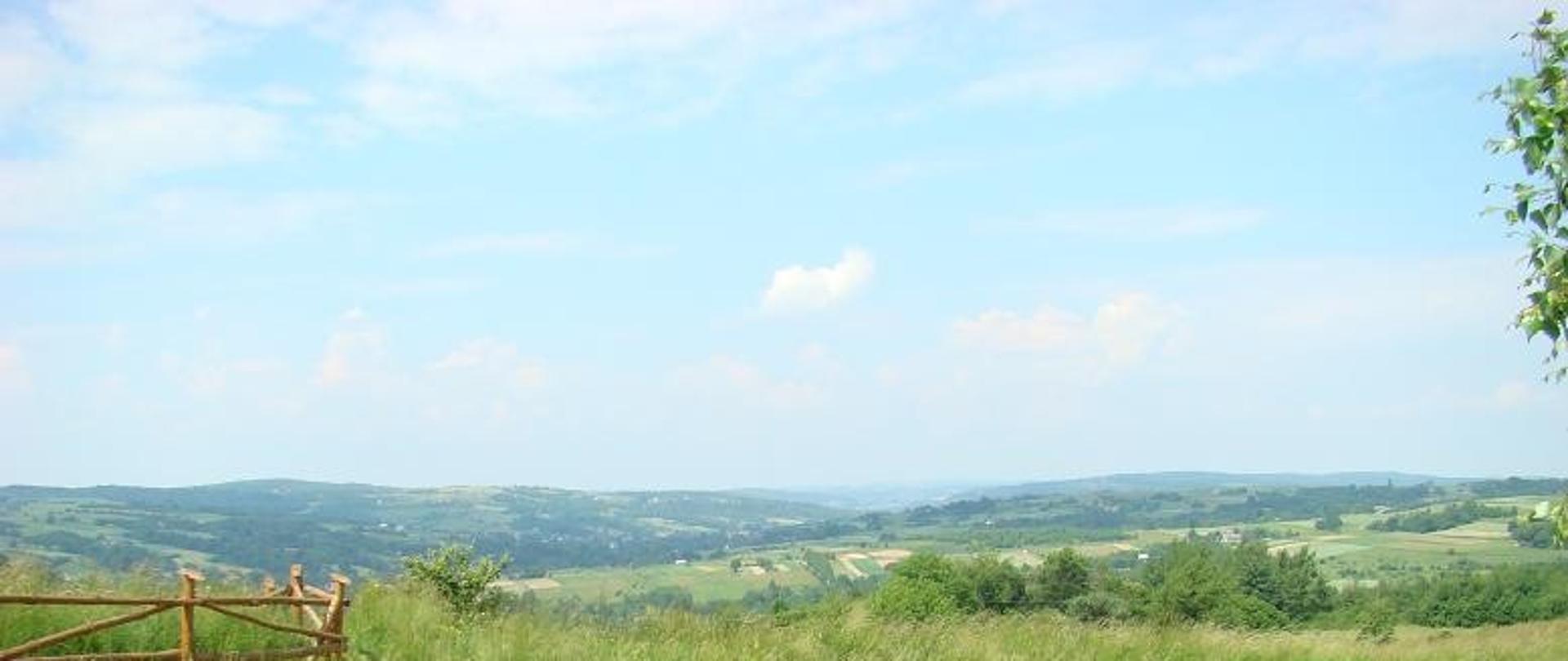 zdjęcie widokowe w oddali miejscowość Lubenia i okolice daleki horyzont widoczne niebo po prawej duży kamień ławka widoczna droga asfaltowa ogrodzenie zielona łąka oraz zadrzewienia