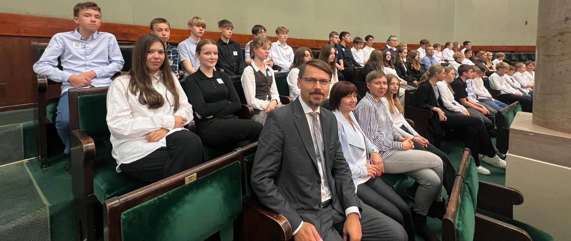 Grupa uczniów oraz mężczyzna siedzą w zielonych ławach beżowego pomieszczenia.