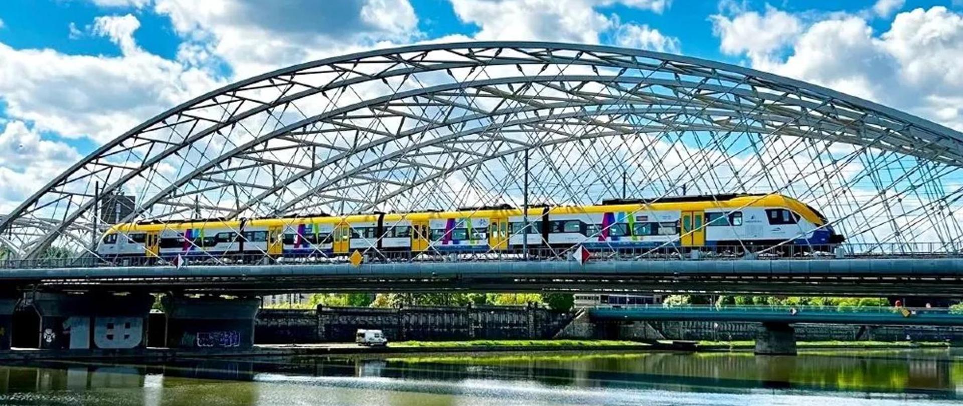Żółto-niebieski pociąg pasażerski jadący po torach na nowoczesnym stalowym moście łukowym nad wodą, w słoneczny dzień z błękitnym niebem i chmurami.