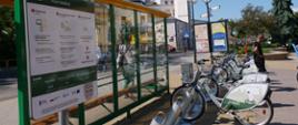 Zdjęcie przedstawia rowery ustawione na stacji roweru miejskiego przy przystanku autobusowym na zewnątrz.