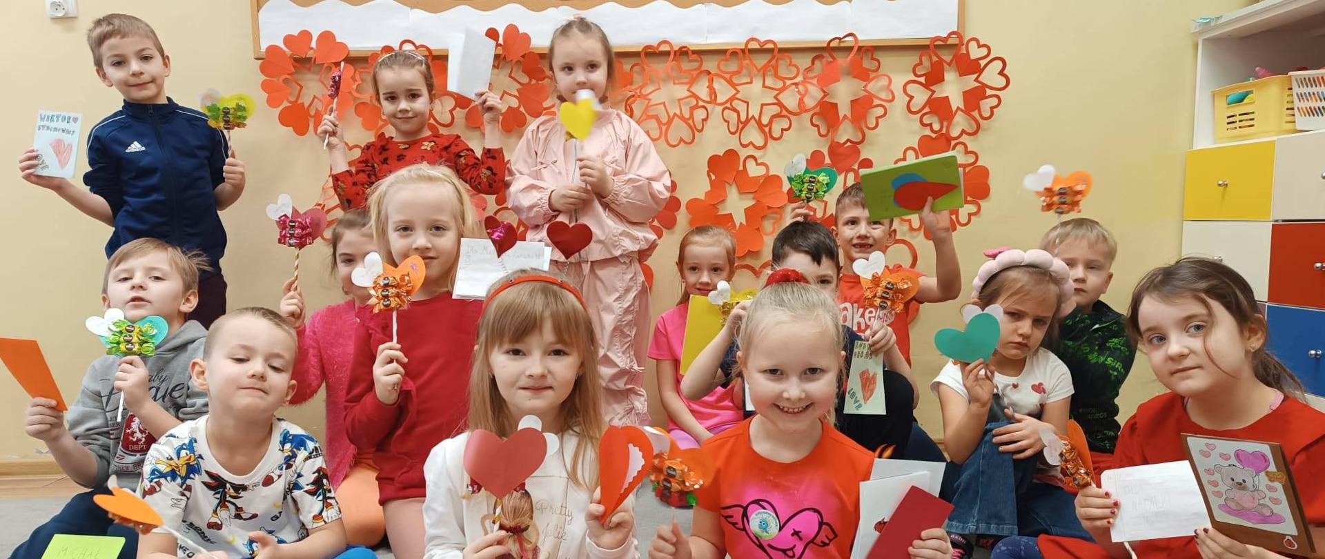 Grupka dzieci w czerwonych strojach siedzi po turecku na dywanie. Wszyscy trzymają serduszka w rękach.