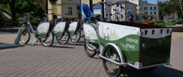 Zdjęcie przedstawia rowery ustawione na kostce brukowej na placu, przy stacji rowerowej. Jeden z rowerów ma przyczepkę z przodu.