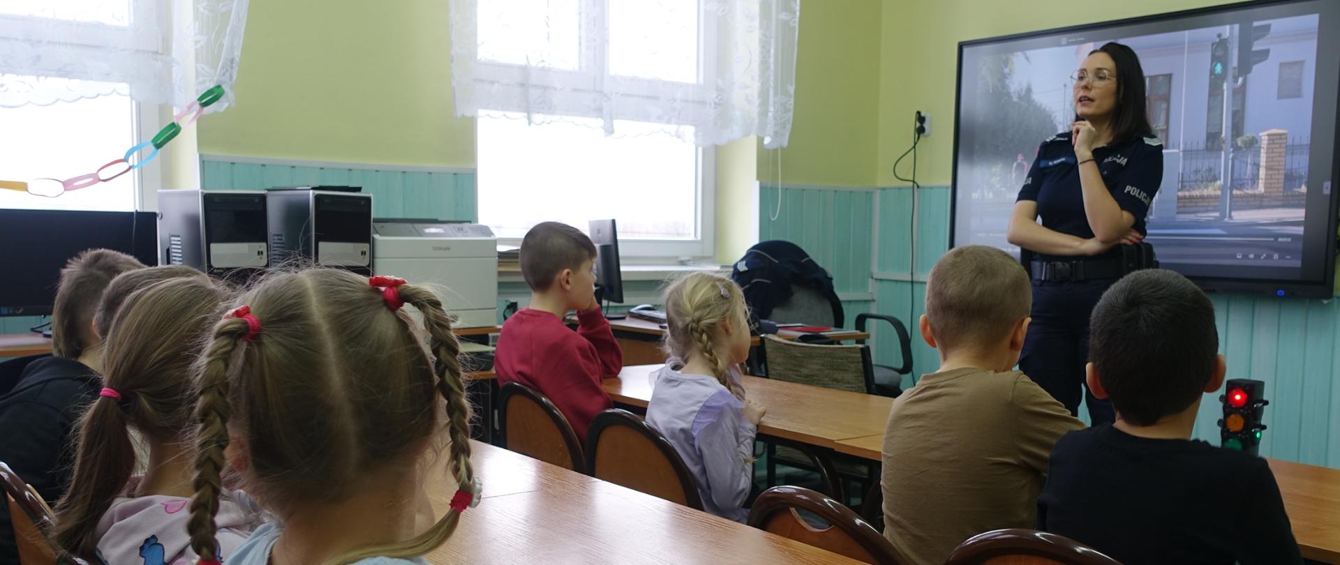 Uczniowie klas młodszych zebrani w sali lekcyjnej uczestniczą w prelekcji Pani policjantki