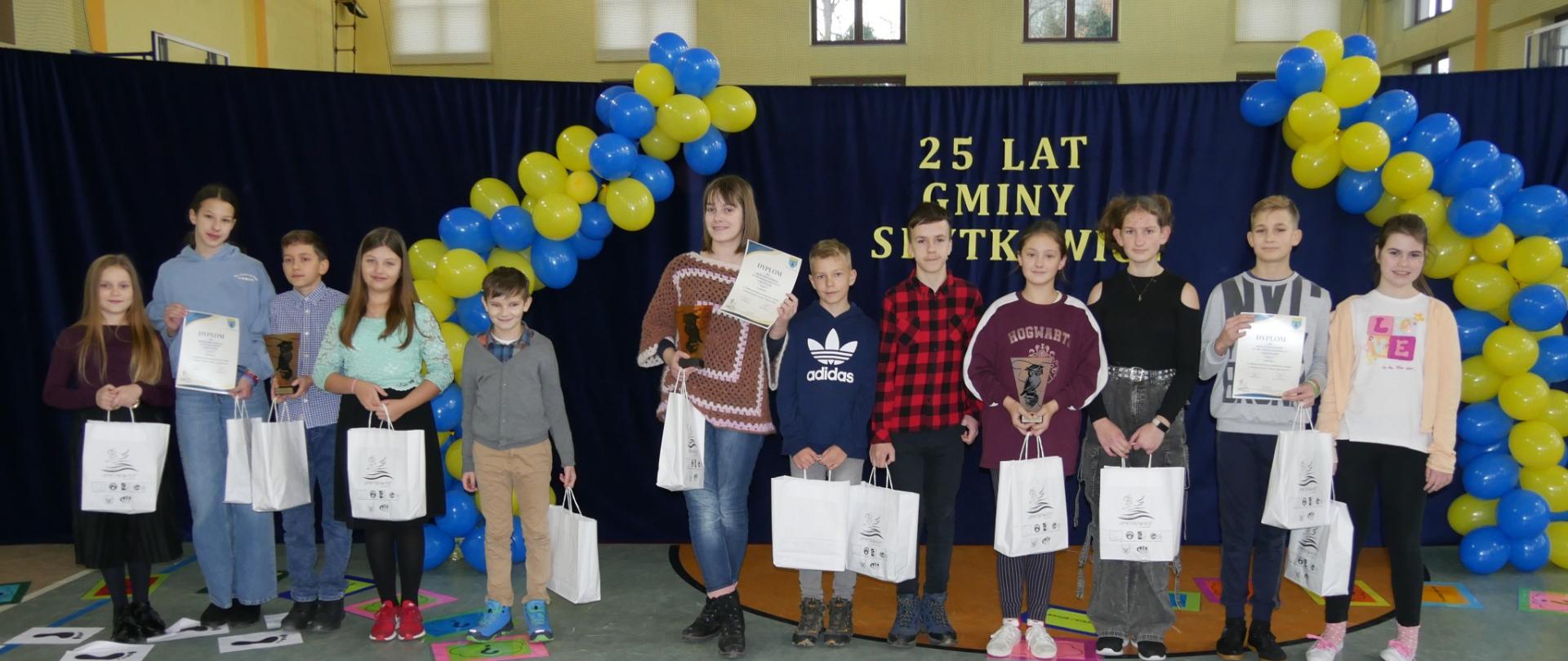 Uczniowie na tle dekoracji z napisem "25 lat Gminy Spytkowice", w tle balony w barwie żółto- niebieskiej, trzymają w papierowe opakowania z nagrodami otrzymanymi w ramach udziału w konkursie.