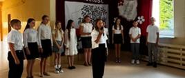 Uczniowie i uczennice z klasy 6 ubrani na galowo występują na akademii, w tle dekoracja z napisem "Pamięć i przyszłość"