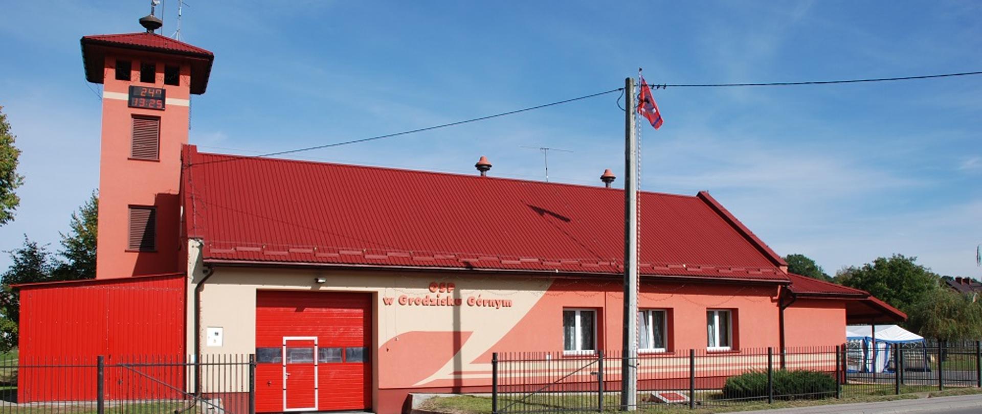 Czerwony, jednopiętrowy budynek z czerwonym, dachem, przy budynku wieża z zegarem i blaszany czerwony garaż. 