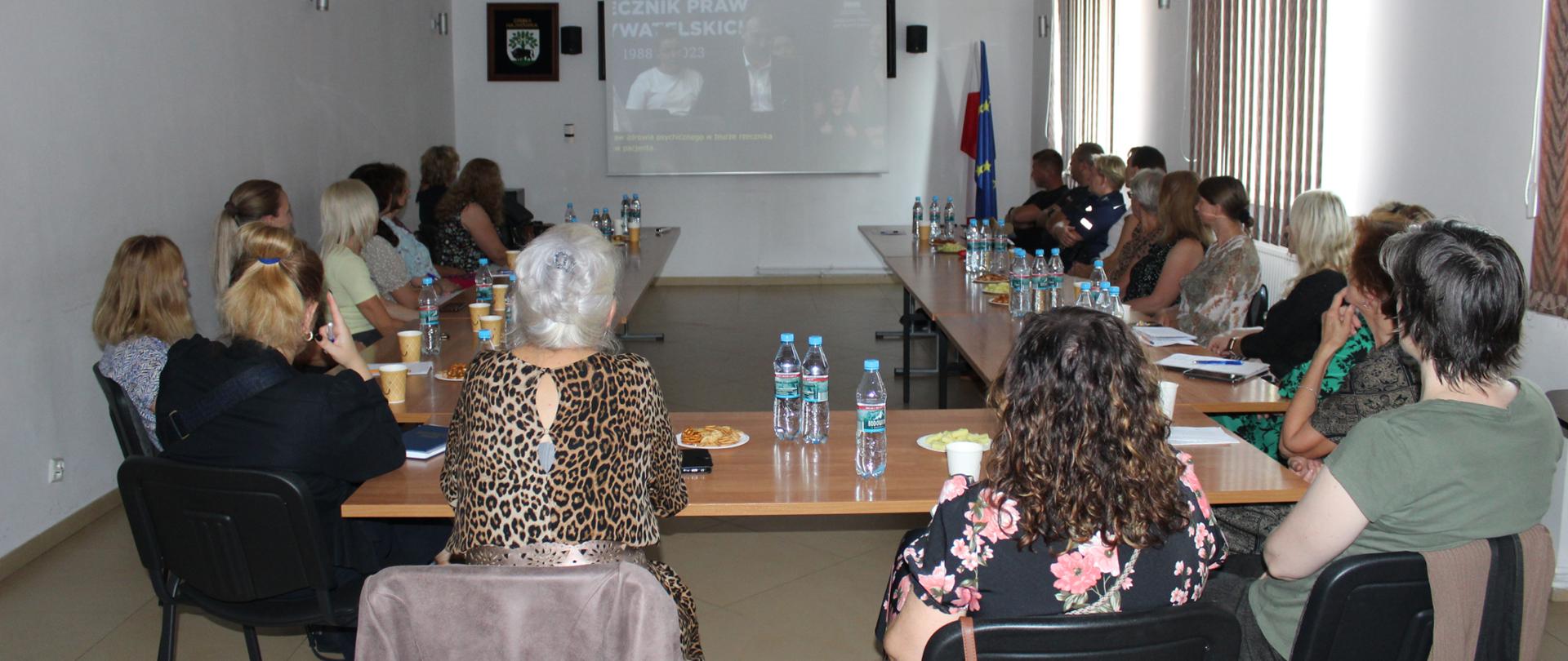 Uczestnicy konferencji siedzą przy biurku, przy monitorze zdalnym