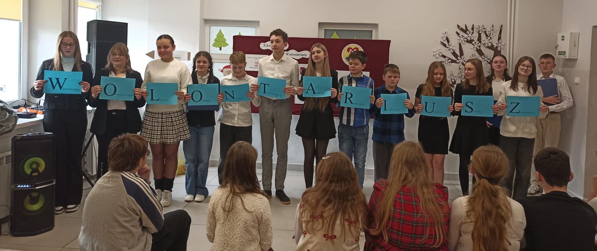 Odświętnie ubrani członkowie Szkolnego Koła Wolontariatu trzymają w ręku - każdy - niebieską kartką z jedną literą słowa "wolontariusz"