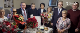 Grupa starszych i dorosłych osób stoi zgromadzona wokół nakrytego stołu w domowym wnętrzu. Na stole znajdują się bukiety kwiatów, tort, ciasta, słodycze, kieliszki i butelka napoju. Kilka osób trzyma bukiety w rękach. Wszyscy pozują do wspólnej fotografii podczas uroczystego spotkania.