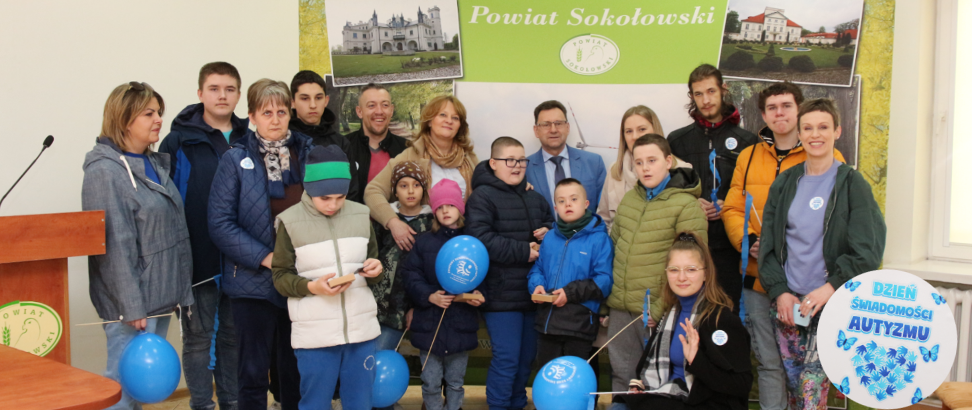 Na fotografii grupa dzieci z Zespołu Szkół Specjalnych im. Jana Pawła II w Sokołowie Podlaskim, która odwiedziła Starostwo z okazji Dnia Świadomości Autyzmu. Dzieci w różnym wieku wraz z dyrektor oraz nauczycielami znajdują się na sali konferencyjnej, pozują do zdjęcia grupowego, niektórzy trzymają w ręku balony i wstążki w kolorze niebieskim. W tle ścianka z fotografiami budynków i napis powiat sokołowski. W prawym dolnym rogu zdjęcia biała okrągła naklejka z niebieskim napisem Dzień świadomości autyzmu, rączek ułożonych w kształcie serca oraz motyli.