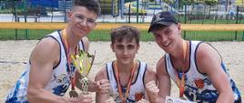 Trzyosobowa reprezentacja chłopców z Zespołu Szkół nr 1 w Kraśniku stojąca na boisku do siatkówki plażowej, prezentująca zwycięskie medale, puchar i dyplom