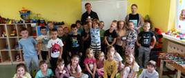 Grupowe zdjęcie dzieci przedszkolnych z dwoma opiekunami. Dzieci stoją i siedzą na kolorowym dywanie z motylami, w sali zabaw z żółtymi ścianami i regałami z zabawkami.
