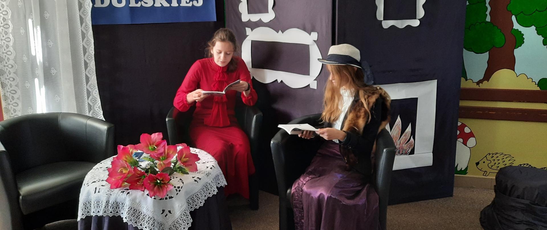 Dwie dziewczyny siedzą na fotelach i czytają książki. Między nimi stoi przykryty haftowanym obrusem stolik. Za nimi na ściance napis Moralność Pani Dulskiej