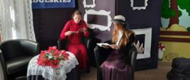 Dwie dziewczyny siedzą na fotelach i czytają książki. Między nimi stoi przykryty haftowanym obrusem stolik. 