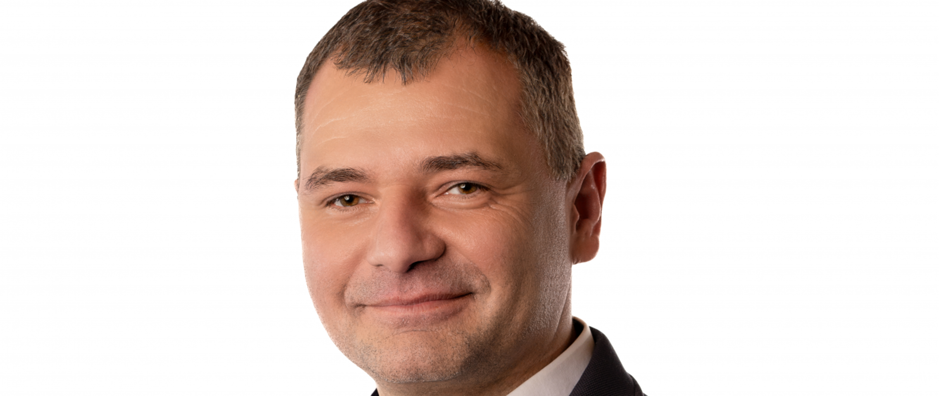 Mężczyzna o ciemnych lekko szpakowatych włosach, małe ciemne oczy, uśmiechnięta twarz. Ubrany w granatowa marynarkę i białą koszule
