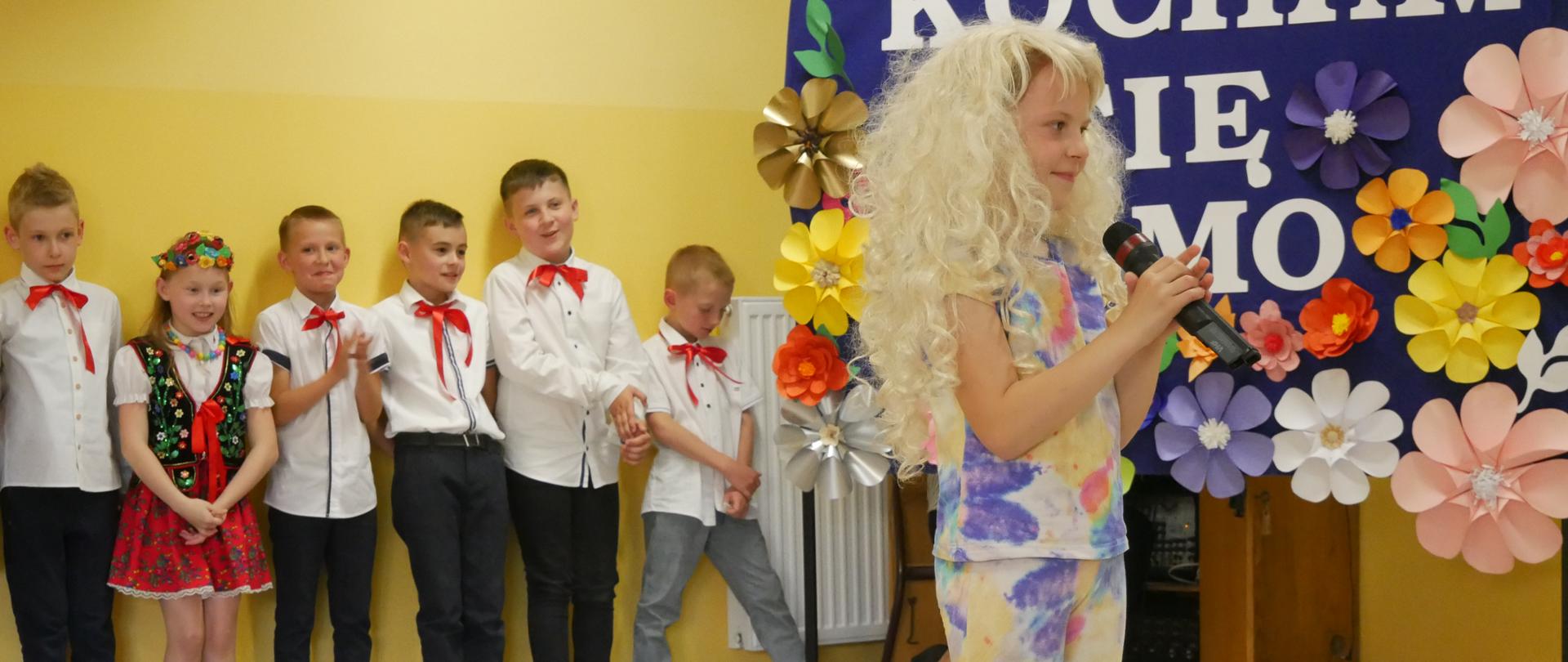Dziewczynka śpiewa piosenkę Majki Jeżowskiej. W tle widać dekorację z kwiatów oraz stojące dzieci: dziewczynki w krakowskich strojach i elegancko ubranych chłopców.
