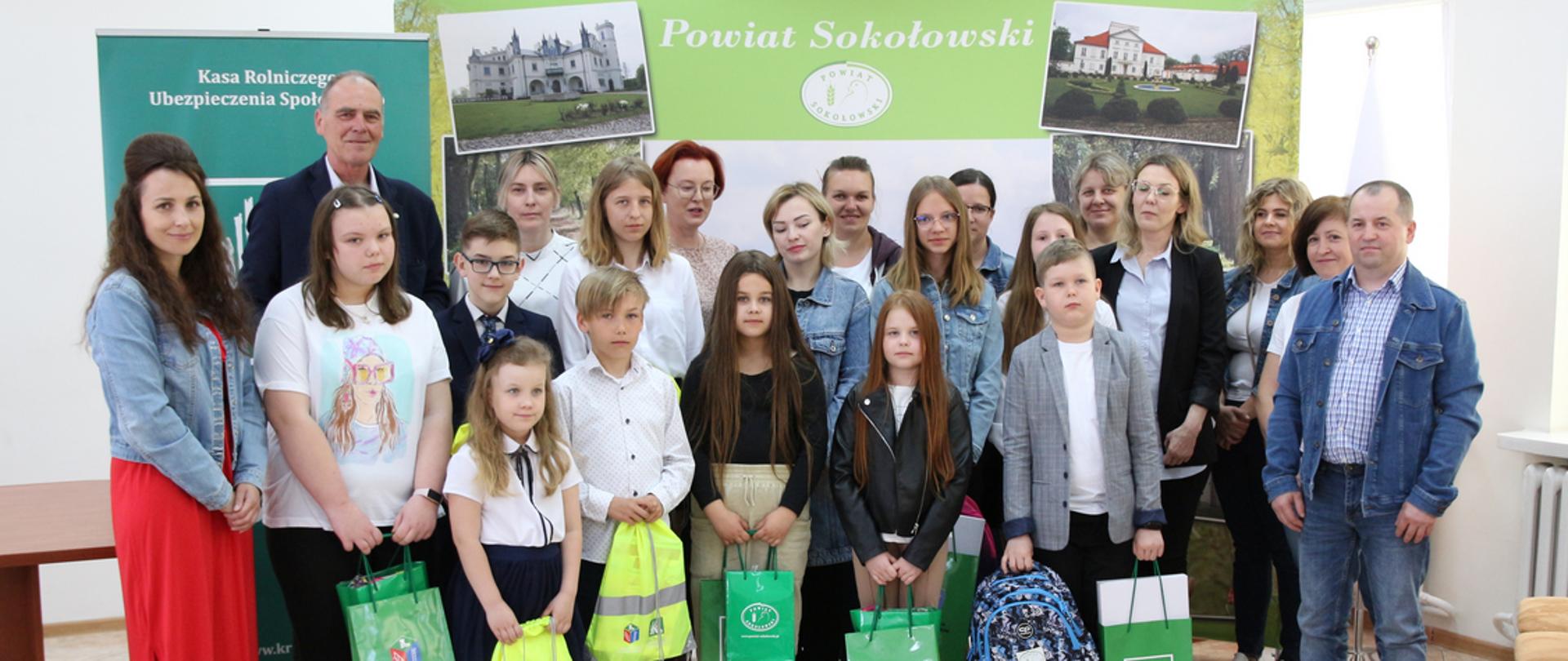 Zdjęcie grupowe laureatów konkursu z organizatorami oraz rodzicami i opiekunami. Dzieci i młodzież wraz z pozostałymi osobami obecnymi podczas uroczystości pozują do fotografii ma tle ścianki, na której widoczny jest logotyp oraz napis powiat sokołowski. Dzieci w dłoniach trzymają zdobyte w konkursie nagrody.