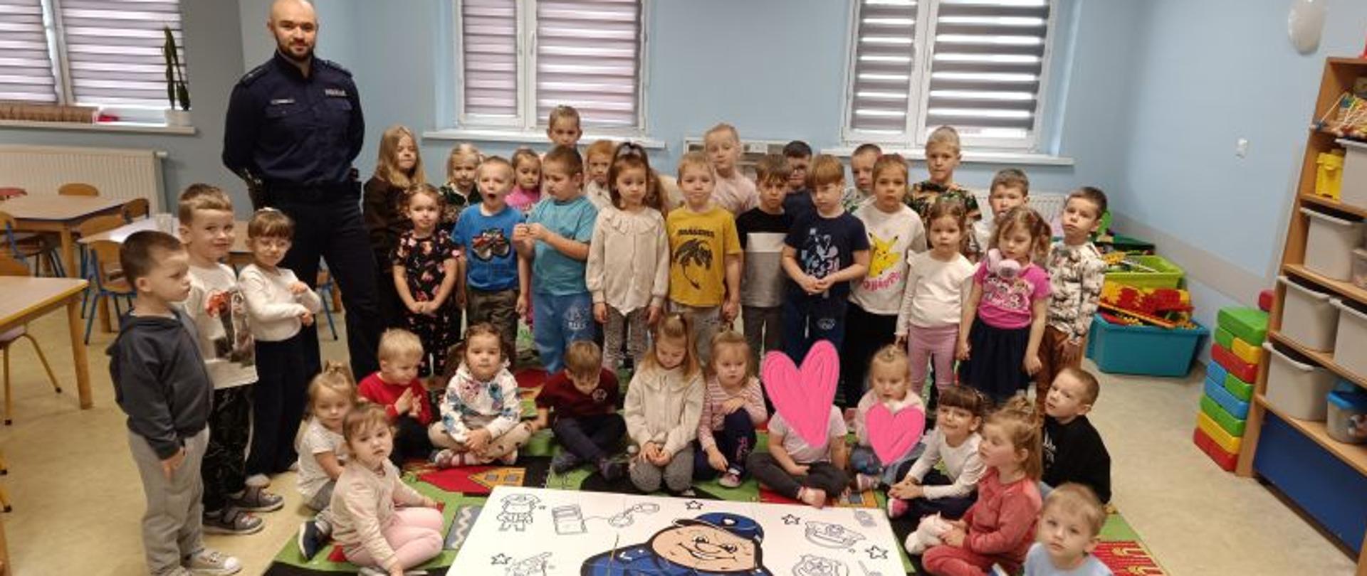 Grupa dzieci, niektóre stoją, inne siedzą. Obok stoi policjant. Przed nimi na dywanie leżą ułożone duże puzzle z postacią Pyrka. 