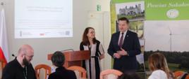 Na fotografii widzimy wicestarostę sokołowskiego stojącego na tle ścianki promocyjnej Powiatu Sokołowskiego wraz z osobą prowadzącą spotkanie. Wicestarosta wita uczestników, których widzimy w pierwszym planie siedzących przy stole. Na Bocznej ścianie wyświetlony jest slajd zatytułowany "Lokalny Plan Rozwoju Usług Społecznych".