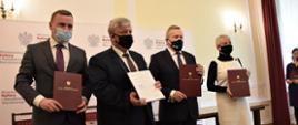 Troje mężczyzn w garniturach oraz kobieta trzymają w rękach przed sobą teczki z orłem
