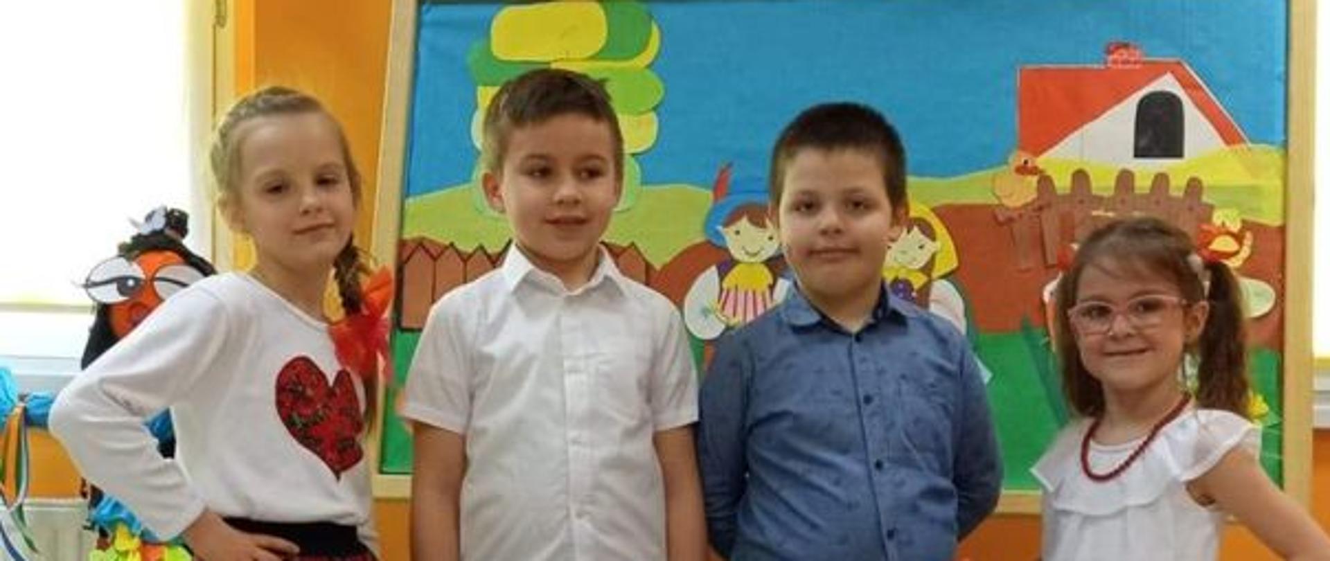 Trzy dziewczynki klęczą, jedna z nich ma wianek na głowie, za nimi stoją dwie dziewczynki i dwóch chłopców. Dziewczynki mają na sobie elementy stroju ludowego, chłopcy eleganckie koszule i czarne spodnie. Za nimi dwa okna a między nimi wisi godło Polski.