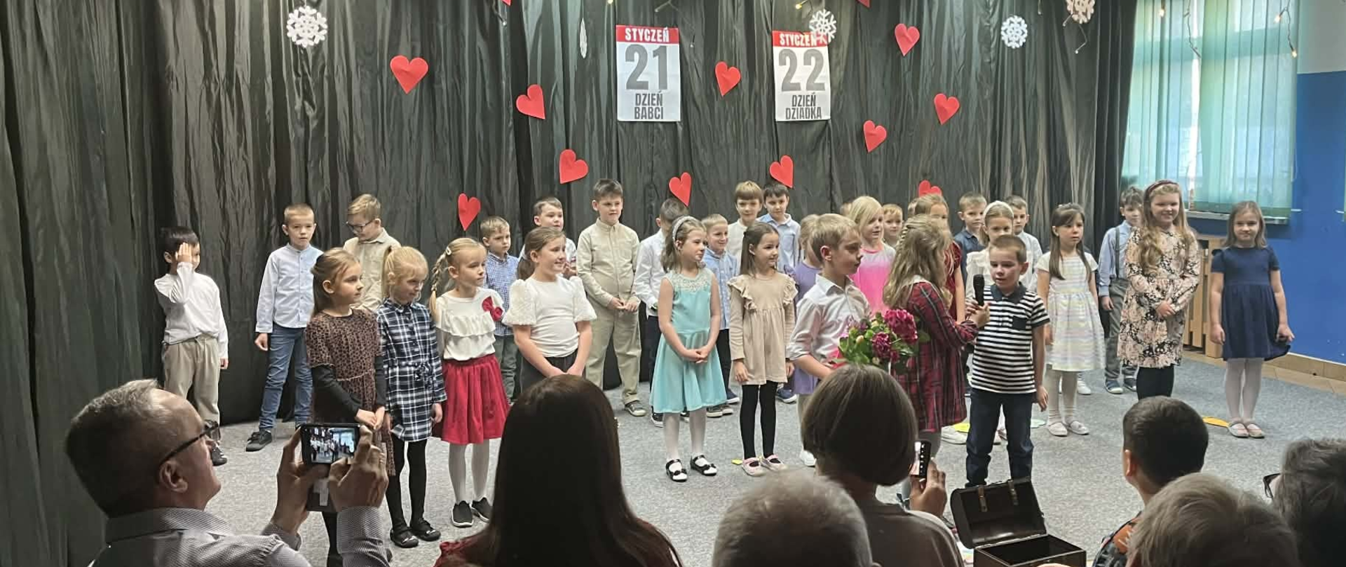 Dzieci stojące w dwóch rzędach. Za nimi ściana pokryta ciemną tkaniną. Na niej czerwone serca oraz liczby 21 i 22. Przed dziećmi dorośli jako widownia.