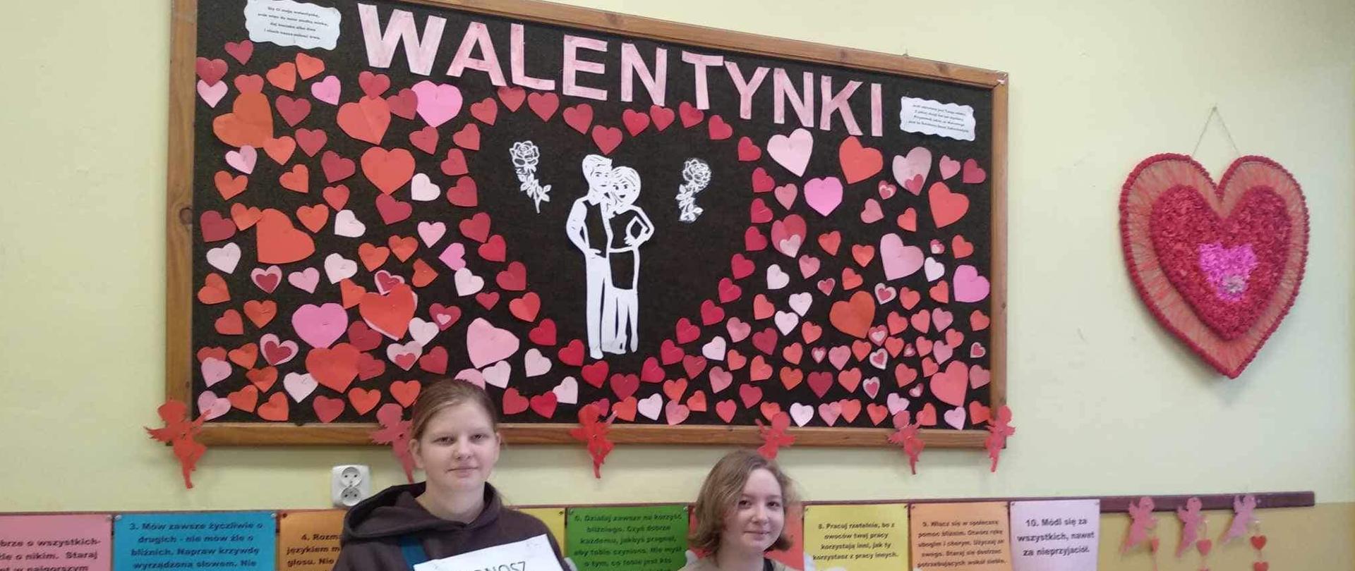 Dwie listonoszki z walentynkowym pudełkiem zawierającym listy i plakatem pozują pod gazetką ścienną informującą o walentynkach