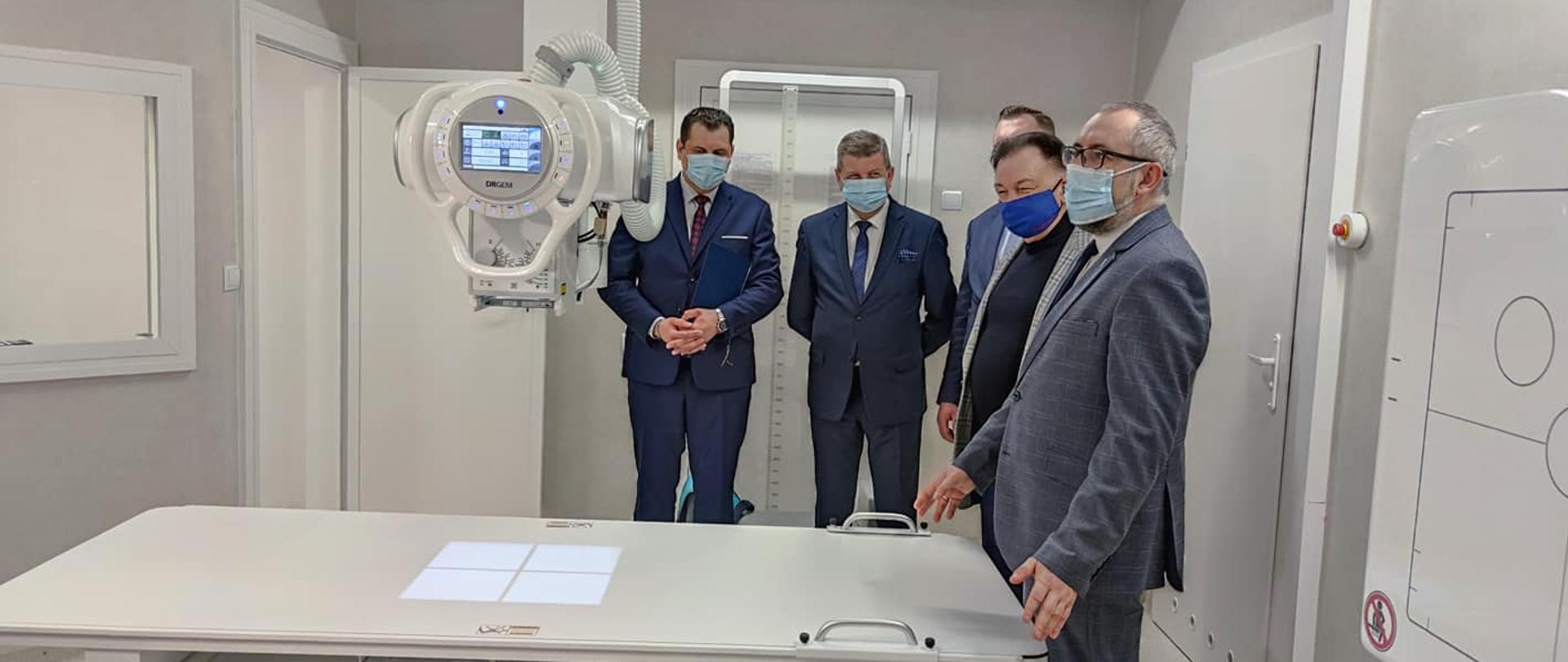Zdjęcie przedstawia wnętrze nowej pracowni rentgenowskiej chwilę po oficjalnym otwarciu. Marszałek Województwa Mazowieckiego Adam Struzik, Radny Sejmiku Woj­ewództwa Mirosław Au­gustyniak, Dyrektor Szpitala Grzegorz Magnuszewski oraz Starosta Przasnyski Krzysztof Bieńkowski stoją przy nowej aparaturze.