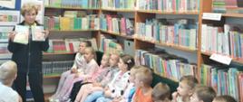 Uczniowie klasy 1 siedzą na ławeczkach w bibliotece, słuchają pani bibliotekarki, która opowiada o autorce :Szewczyka Dratewki" pisarce Janinie Porazińskiej i trzyma w rękach 2 książki autorki 