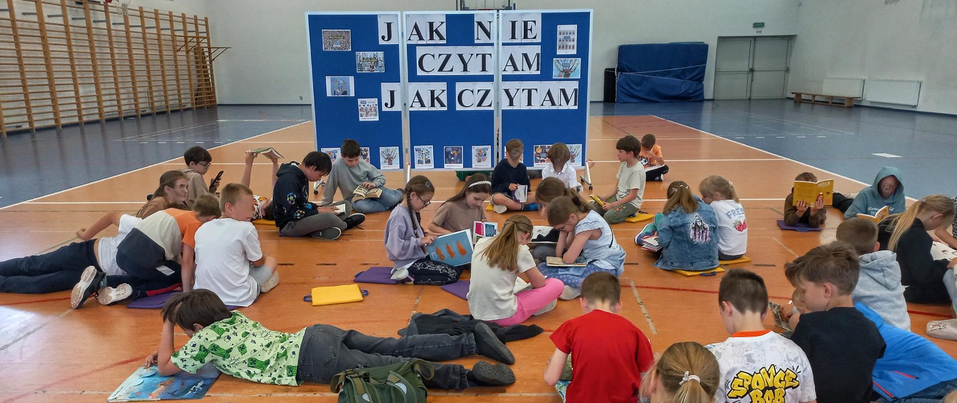 Dzieci siedzące z książkami na podłodze na sali gimnastycznej. Za nimi widać niebieską tablicę z dużym biało - czarnym napisem. Po prawej stronie widać drabinki gimnastyczne, a po lewej ławki pod ścianą,
