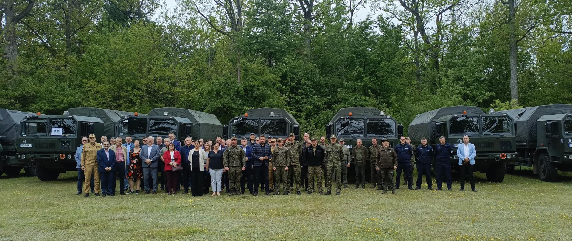 Uczestnicy spotkania pozują do zdjęcia na tle floty aut