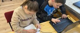Przy ławce szkolnej siedzą uczennica - od lewej - i uczeń. Dziewczynka pisze na kartce długopisem 3D, chłopiec pisze na klawiaturze laptopa