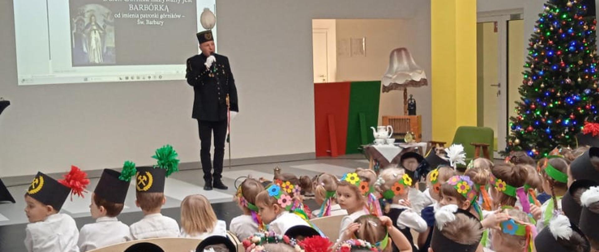 Górnik stoi w jasnej sali przed grupą siedzących przedszkolaków