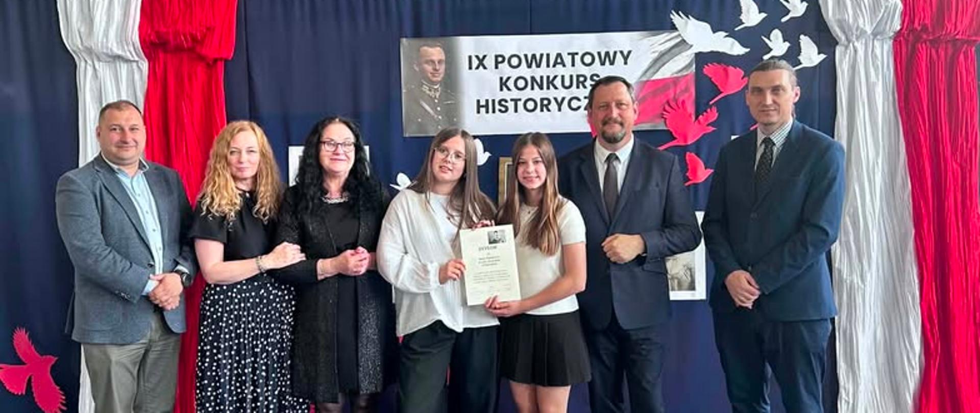 Na zdjęciu widać grupę siedmiu osób stojących na sali gimnastycznej podczas uroczystości związanej z konkursem historycznym. W centrum znajdują się dwie uczennice – jedna z nich trzyma dyplom, co sugeruje, że zostały nagrodzone lub wyróżnione za swoje osiągnięcia.
Tłem jest dekoracja w barwach biało-czerwonych z napisem „IX Powiatowy Konkurs Historyczny”. Obok napisu znajdują się sylwetki lecących ptaków oraz elementy nawiązujące do historii i patriotyzmu. Nad dekoracją wiszą banery związane z wydarzeniami historycznymi, m.in. biegiem pamięci.
Po obu stronach uczennic stoją dorośli – prawdopodobnie nauczyciele, opiekunowie, organizatorzy konkursu lub przedstawiciele władz oświatowych. Wszyscy są ubrani elegancko, co podkreśla oficjalny charakter wydarzenia. Osoby na zdjęciu uśmiechają się i pozują do pamiątkowej fotografii po wręczeniu nagrody.