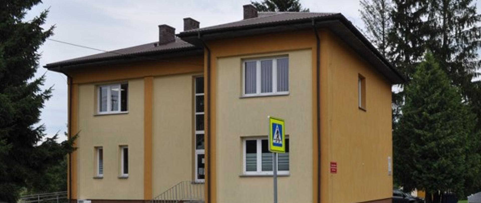 Na zdjęciu widoczny budynek komunalny w którym znajduje się Gminny Ośrodek Pomocy Społecznej w Brańszczyku. Budynek ma wykonaną elewację w kolorze pomarańczowym oraz brązowy dach. Wokół budynku rosną wysokie drzewa iglaste, a działka ogrodzona jest szarym metalowym płotem.