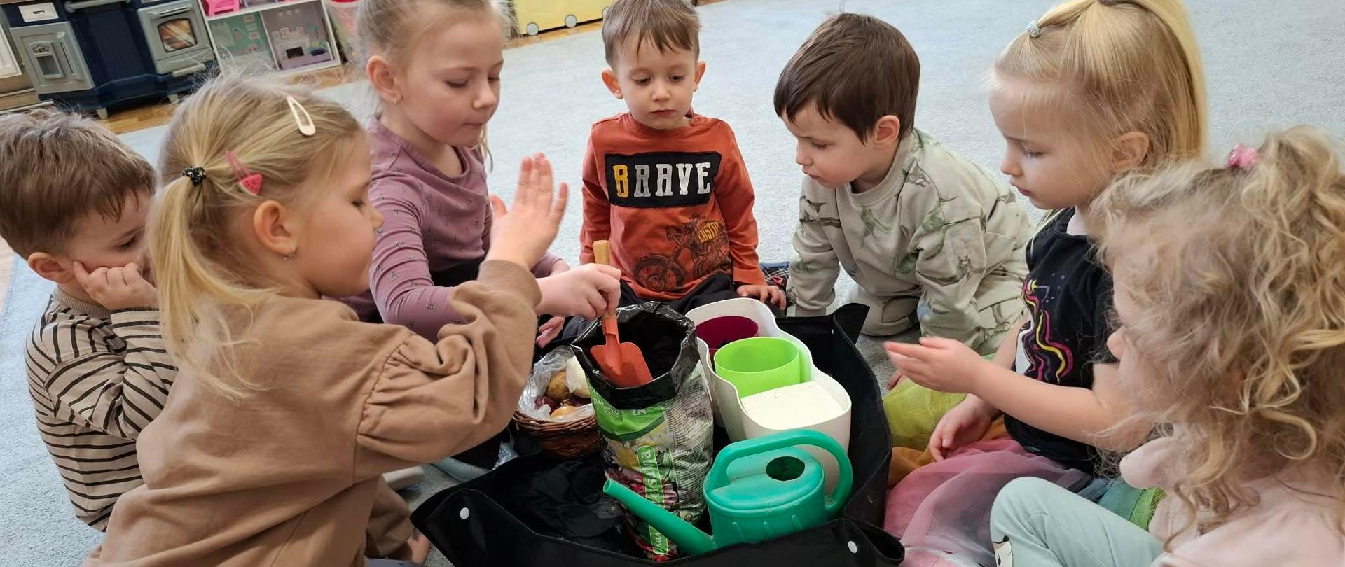 Grupa dzieci siedzi po turecku w kółeczku na dywanie a po środku nich konewka, worek ziemi ogrodniczej, doniczki i koszyk z cebulkami kwiatów.
