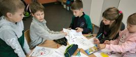Troje chłopców i 2 dziewczynki siedzą przy stoliku na którym są porozkładane kartki z kolorowankami. Dzieci kolorują i przekładają kartki