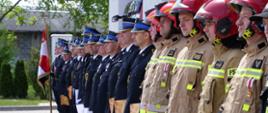 Zdjęcie przedstawia kilkadziesiąt osób stojących w rzędzie. Część ma na sobie galowe mundury strażackie, pozostali typowy strój roboczy.