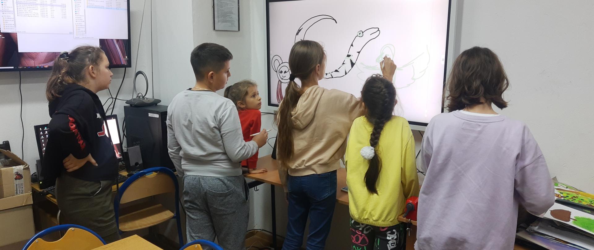 Na zdjęciu dzieci z Ukrainy rysują na nowym monitorze interaktywnym, dziewczynka z długimi włosami spiętymi w kucyk ma w dłoni specjalny rysik, pozostałe dzieci z zainteresowaniem przyglądają się procesowi tworzenia w nowej technologii. 