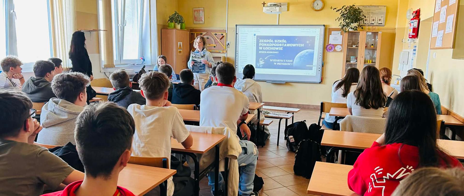 Dzieci siedzące w sali przy ławkach szkolnych. Przed nimi stoi kobieta i trzyma oburącz przed sobą plakat z wizerunkiem budynku. Na tablicy wyświetlona jest prezentacja z napisem Zespół Szkół Ponadpodstawowych w Łochowie w naszej szkole