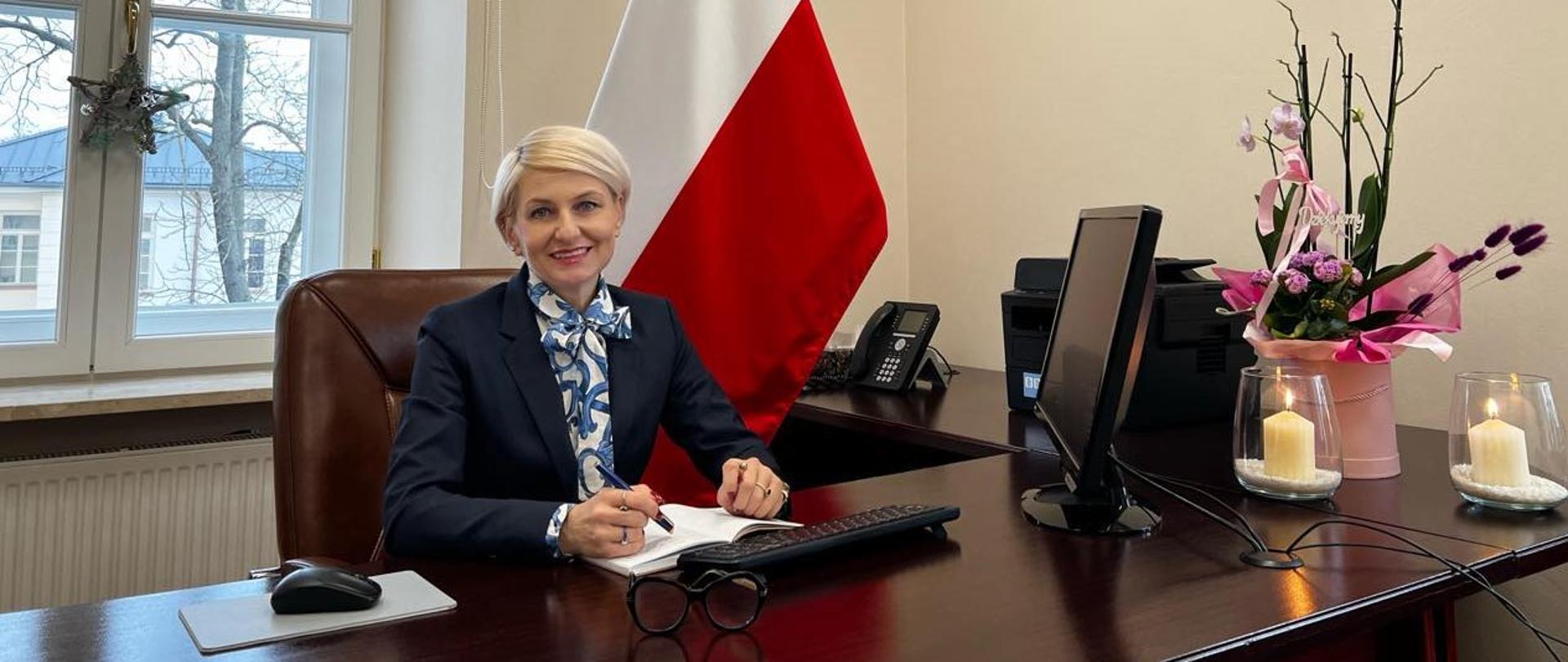 Wicedyrektor szkoły mgr Agnieszka Zdanowska siedząca przy biurku z komputerem na tle biało-czerwonej flagi.