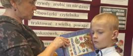 Pani bibliotekarka pasuje ucznia na czytelnika poprzez położenie książki na jego ramieniu
