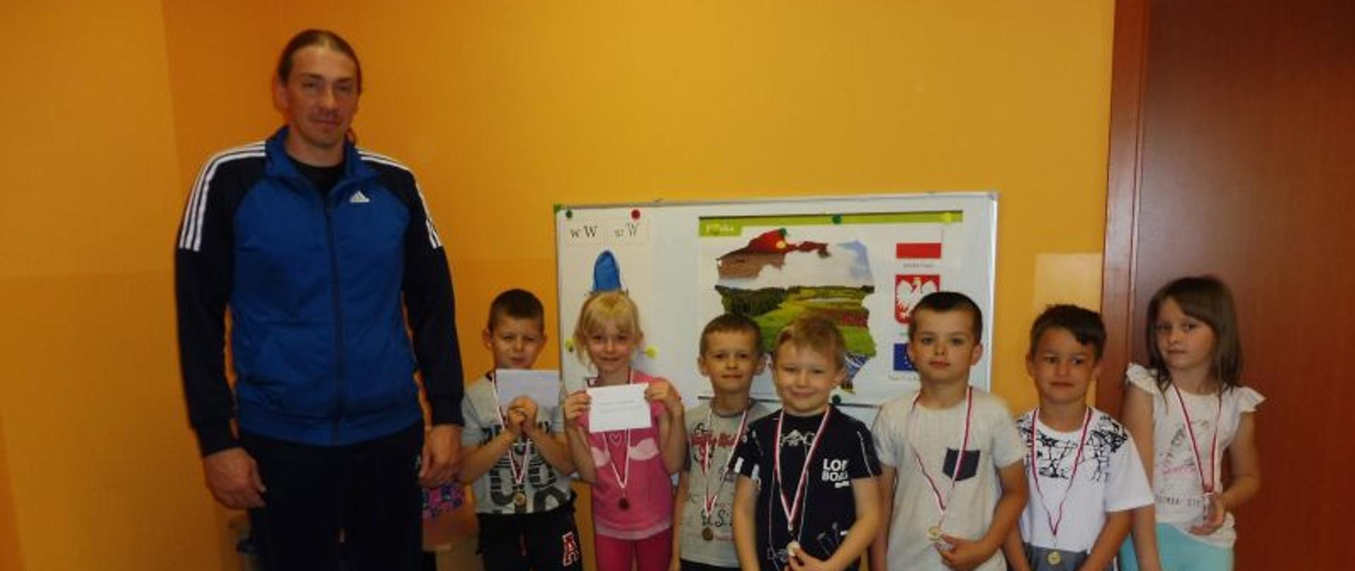 Grupa dzieci, która wzięła udział w turnieju warcabowym, na szyjach mają powieszone medale, obok stoi mężczyzna.