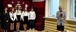 Uczniowie klasy ósmej ubrani na galowo prezentują program artystyczny na korytarzu szkolnym, w tle na bordowej zasłonie dekoracja związana z okazją: biało-czerwona flaga oraz wycięte z papieru: otwarta książka, czapka absolwenta i pióro, przed uczniami pani dyrektor szkoły przemawia do zgromadzonych dzieci